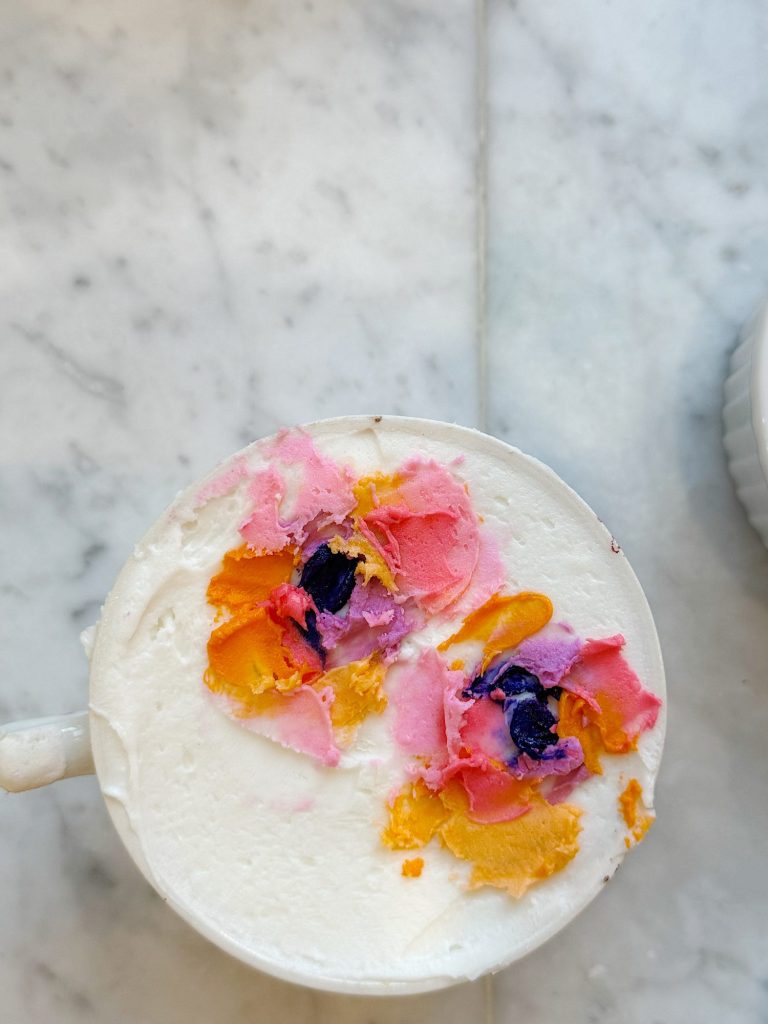 A cup of milk foam decorated with colorful flower patterns in pink, yellow, and purple on a marble surface.