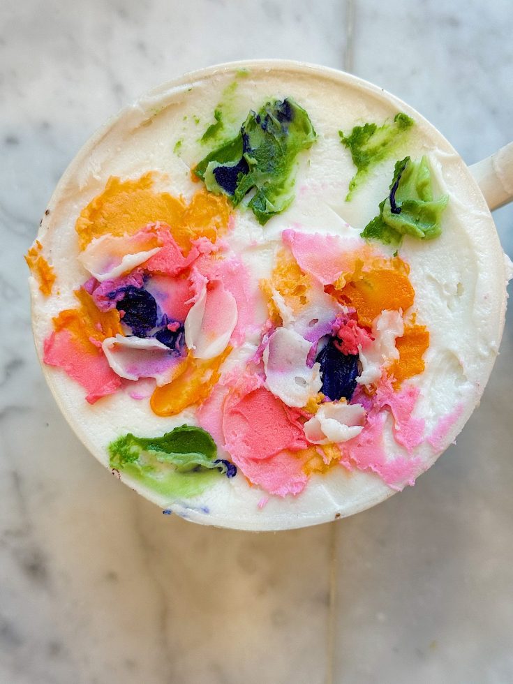 A cup of coffee with steamed milk foam decorated with colorful floral latte art in pink, orange, green, and purple, on a marble surface.