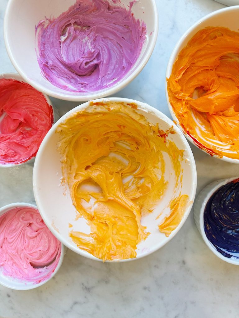 Bowls of colorful frosting in pink, purple, yellow, orange, and dark blue on a marble surface, ready for decorating.