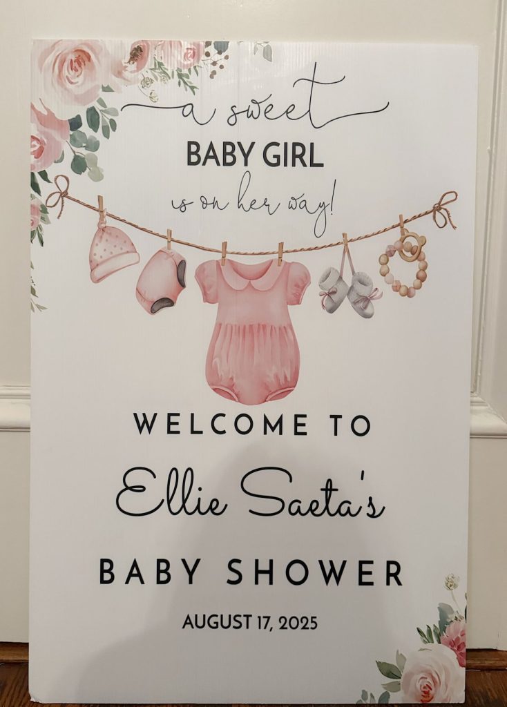 A sign decorated with floral designs and baby clothes announces "Ellie Saeta's Baby Shower" for a baby girl on August 17, 2025.