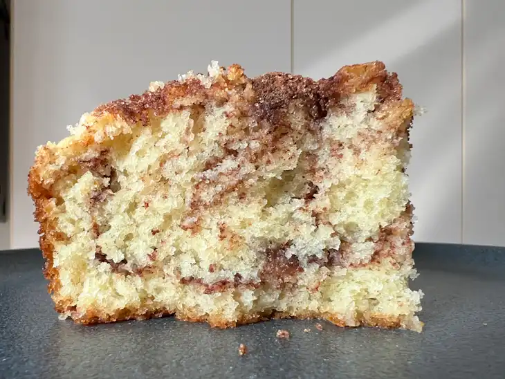 A close-up of a slice of coffee cake with a cinnamon swirl, placed on a dark surface.