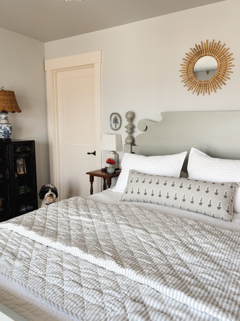 A neatly made bed with patterned bedding, decorative pillows, and a tufted headboard. A small dog sits by the bedside table. A round mirror and lamp are on the wall.