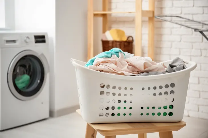 A white laundry basket filled with clothes sits on a wooden stool in a laundry room with a washing machine in the background.