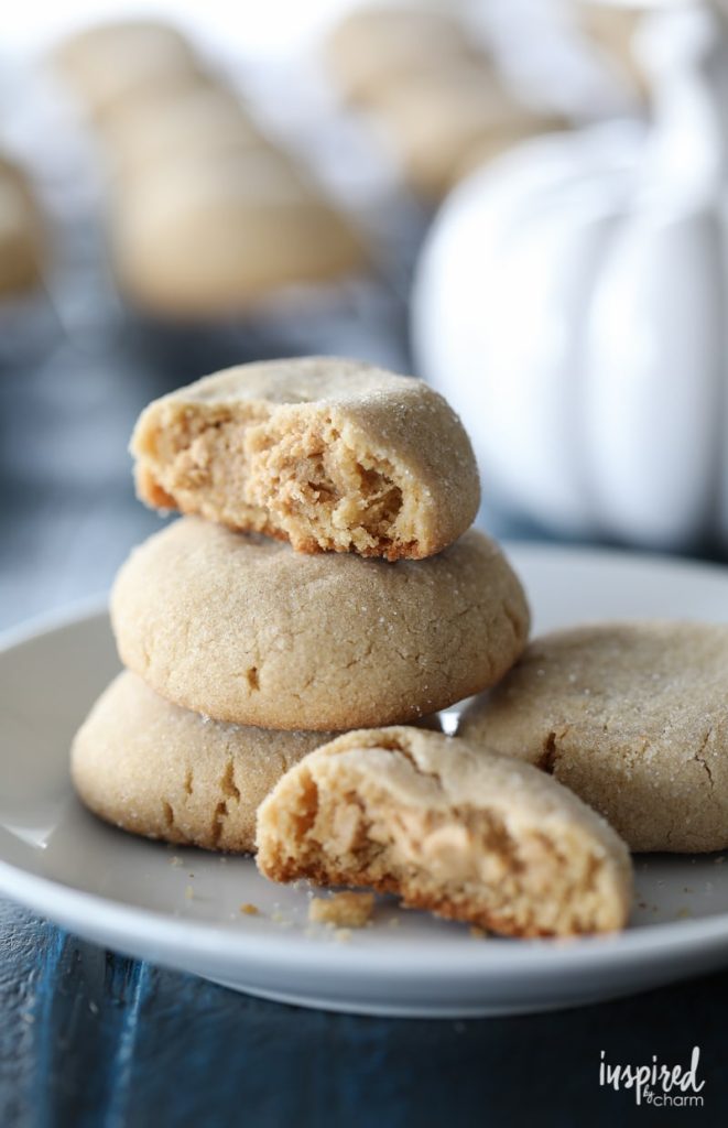 Three sugar-coated cookies are stacked on a white plate, with one broken open to show a soft, crumbly interior. More cookies and a white ceramic pumpkin are blurred in the background.