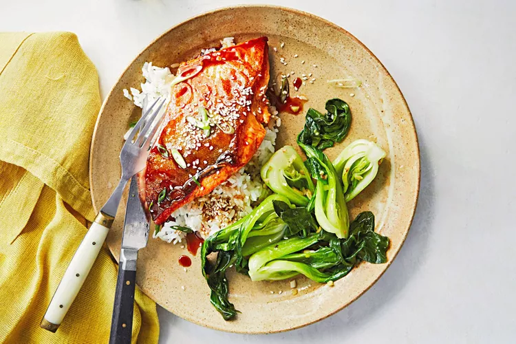 A plate with glazed salmon on rice, sautéed bok choy, and a sprinkle of sesame seeds, with a fork and knife placed on a yellow napkin.