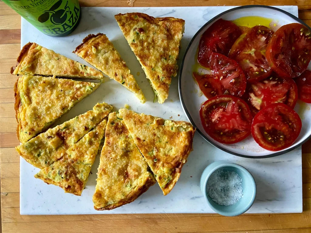 A sliced savory tart on a marble board beside a plate of sliced tomatoes with olive oil and a small bowl of salt.