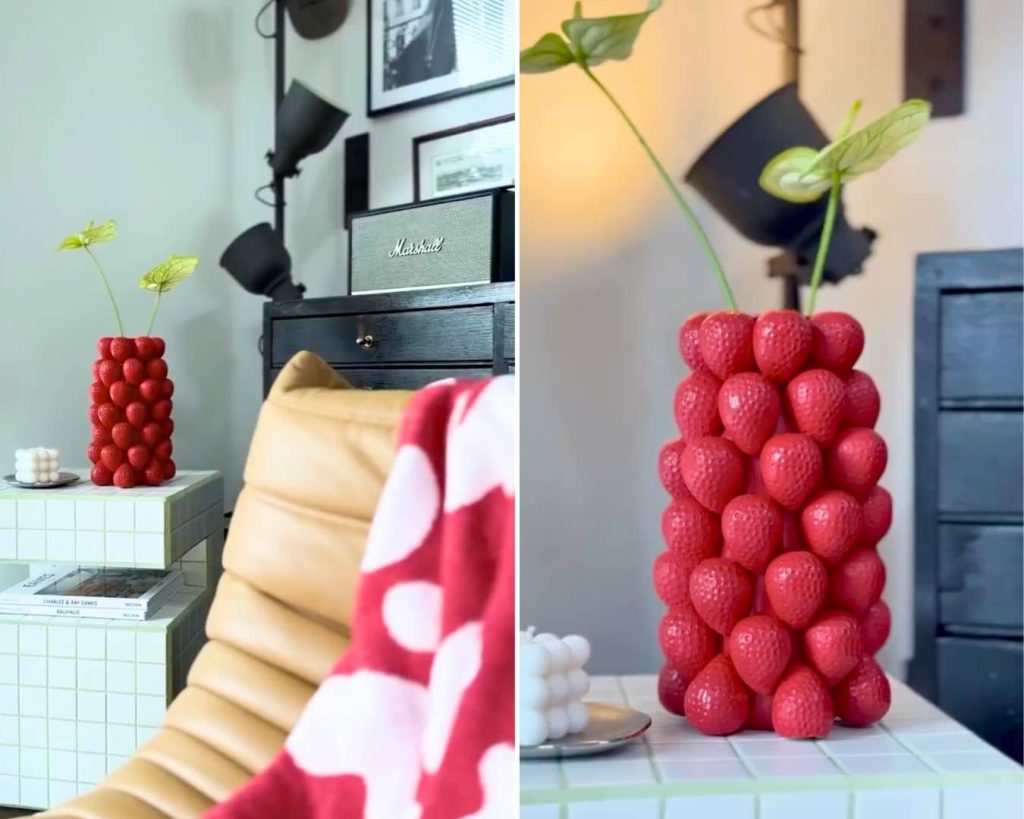 A vase covered with red strawberry shapes holds a couple of green stems. It is placed on a tiled side table next to a tan chair and a red-and-white blanket.