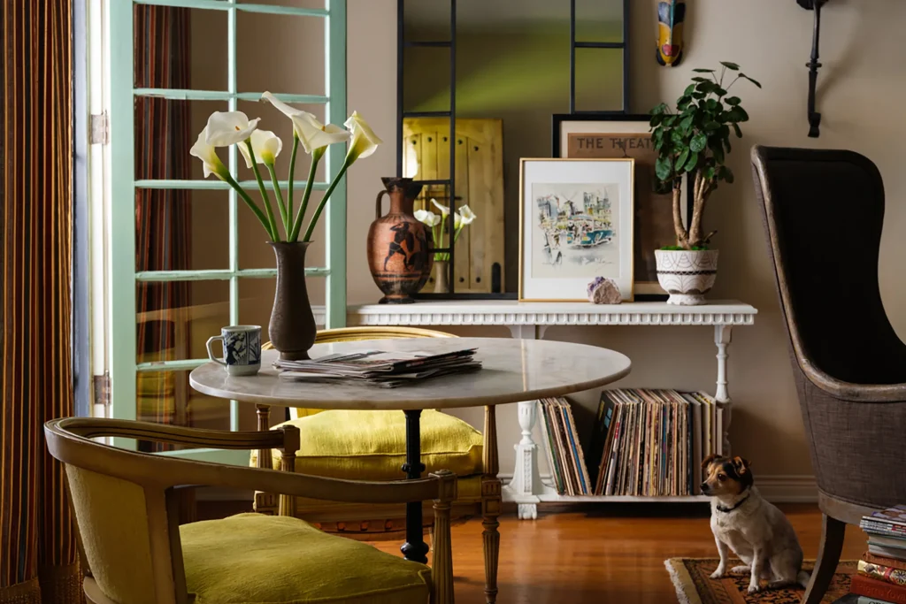 A cozy living room with a round table, yellow-cushioned chairs, vase of white flowers, potted plant, art, vinyl records, and a small dog sitting on the floor.