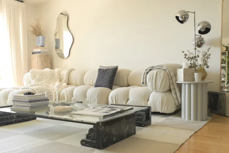 Modern living room with a cream sofa, black marble coffee table, decorative mirror, floor lamp, and neutral decor accents on a striped rug.