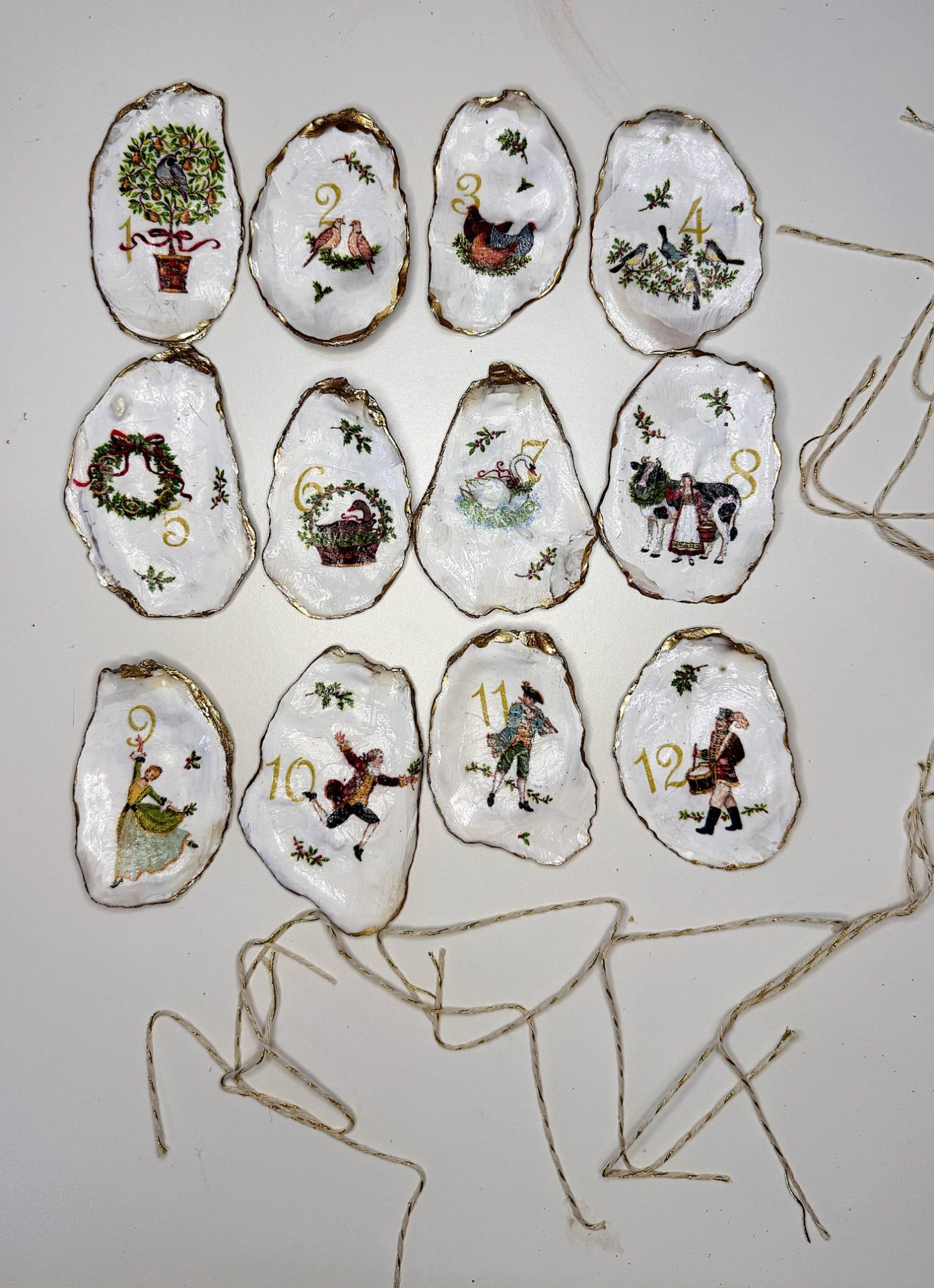 Twelve hand-painted oyster shell ornaments with Christmas-themed illustrations and numbers 1 to 12, each with an attached string for hanging, arranged on a white surface.