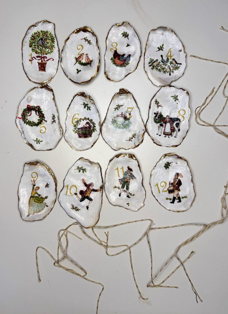 Twelve hand-painted oyster shell ornaments with Christmas-themed illustrations and numbers 1 to 12, each with an attached string for hanging, arranged on a white surface.
