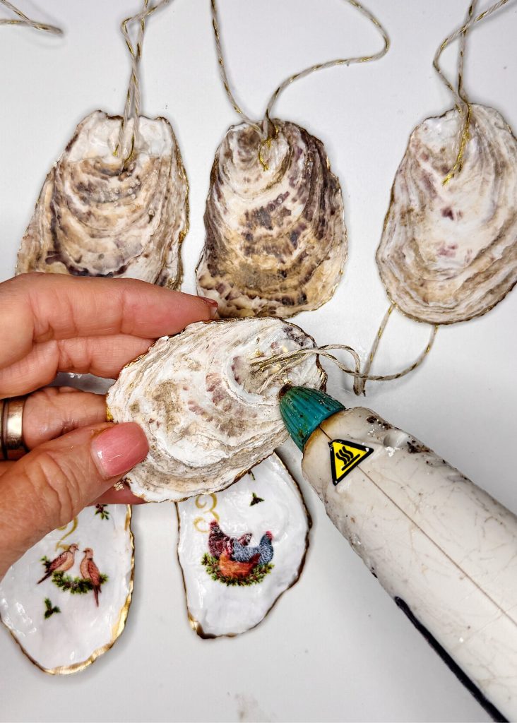 A hand uses a hot glue gun to attach twine to an oyster shell ornament; decorated oyster shells with images and gold trim are shown below.