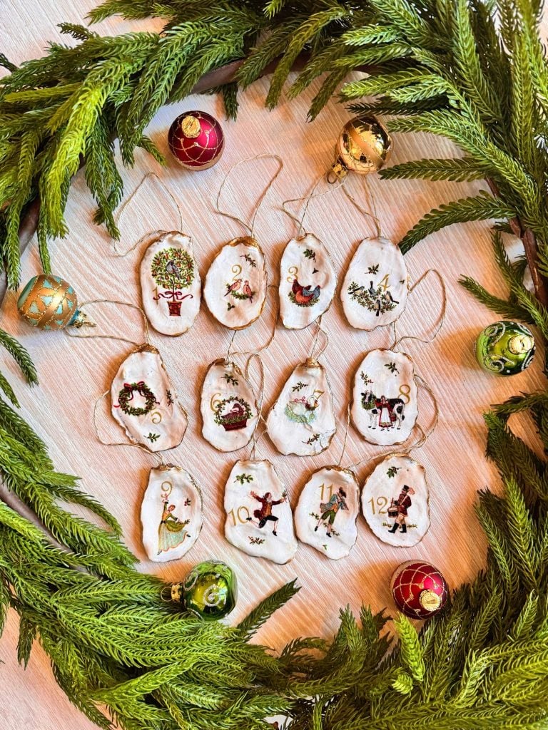 Twelve handmade ornaments painted with images from “The Twelve Days of Christmas” are arranged on a table, surrounded by a green wreath and Christmas baubles.