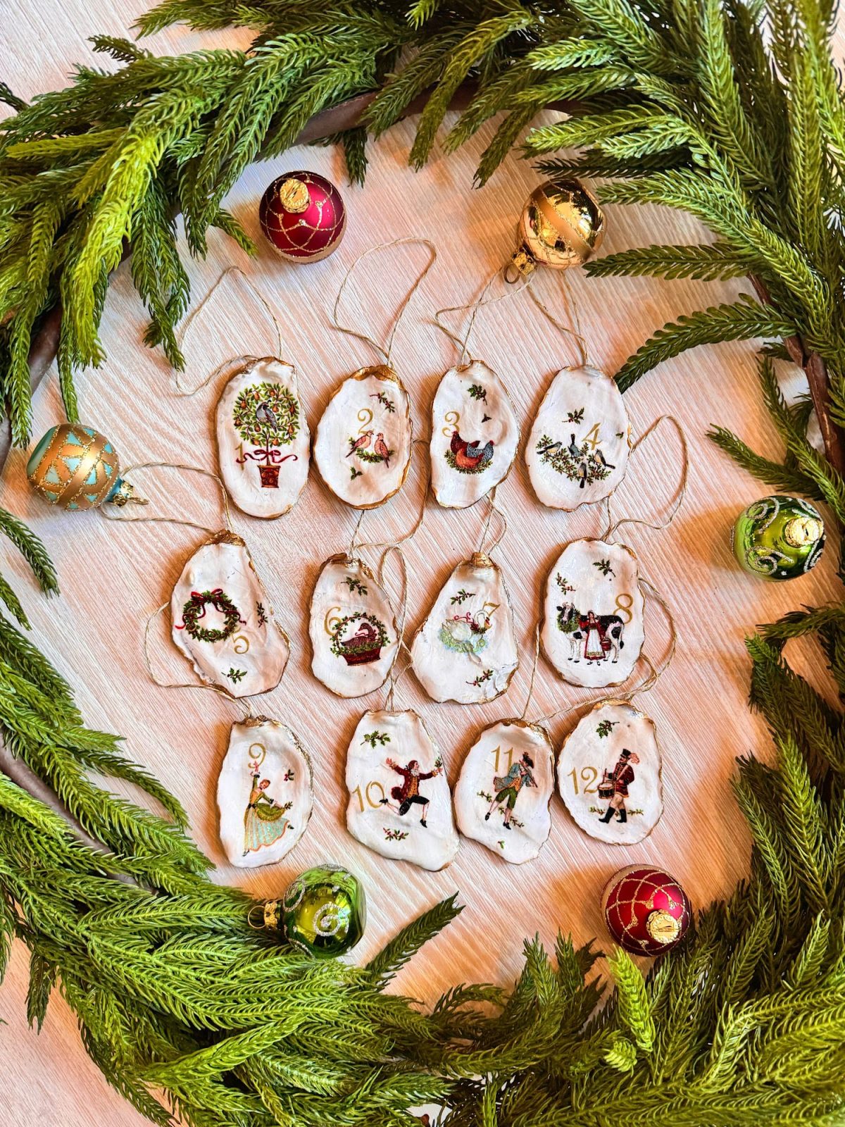 Twelve handmade ornaments painted with images from &ldquo;The Twelve Days of Christmas&rdquo; are arranged on a table, surrounded by a green wreath and Christmas baubles.