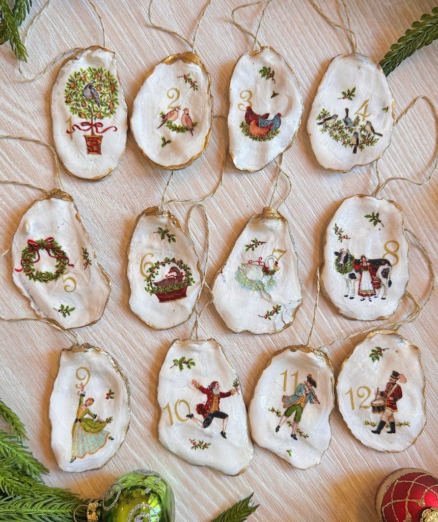 Twelve hand-painted Christmas ornaments on oyster shells, each displaying a number and an illustration from "The Twelve Days of Christmas." Greenery decorates the surrounding area.