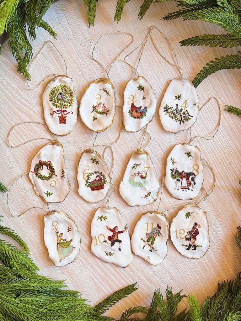 Twelve hand-embroidered fabric ornaments, each depicting a different scene from "The Twelve Days of Christmas," are arranged on a light surface surrounded by greenery.