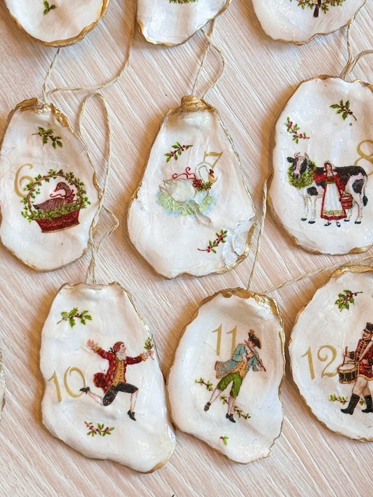 Several oyster shells painted with numbers and festive holiday scenes, each with a gold-rimmed edge and string for hanging, are arranged on a light wooden surface.