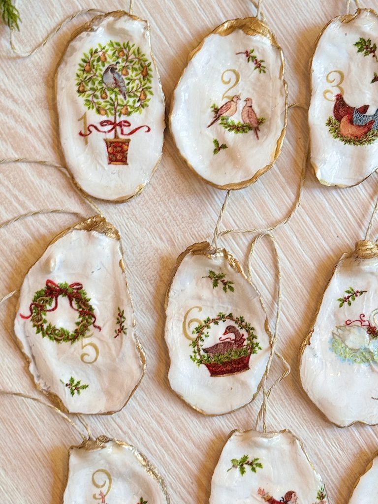 Hand-painted oyster shell ornaments featuring birds, wreaths, and festive designs, each edged in gold and hung with twine, are arranged on a light wood surface.
