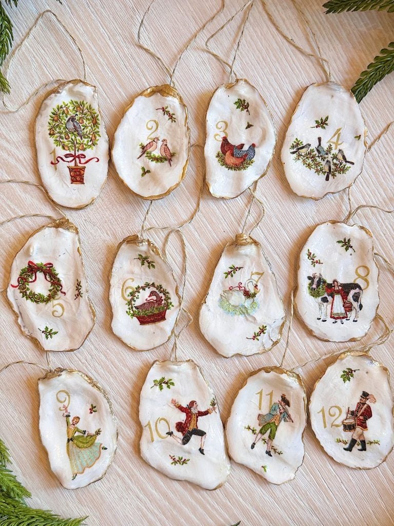 Twelve hand-painted ornaments on oyster shells, each showing a numbered design from "The Twelve Days of Christmas," arranged on a light wood surface.