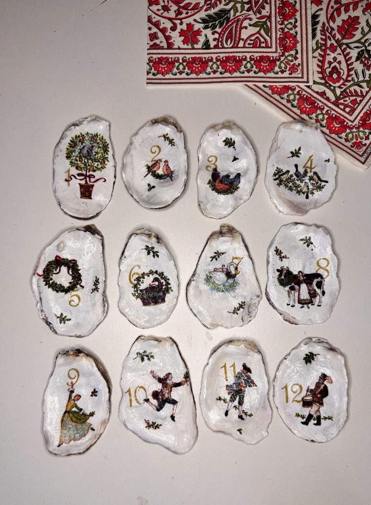 Twelve decorated oyster shells, each painted with a number and a festive image, are arranged in rows on a white surface next to patterned napkins.