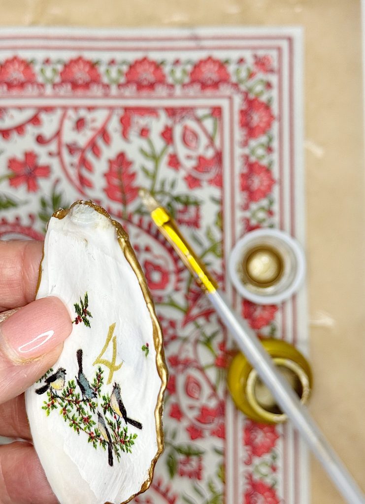 A hand holding a painted oyster shell with a gold rim, number 4, and bird design; a paintbrush and gold paint are on a patterned surface in the background.