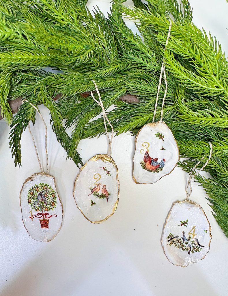 Four oval ornaments with painted holiday designs hang on string in front of green pine branches. Each ornament has a gold-trimmed edge and features a different festive illustration.