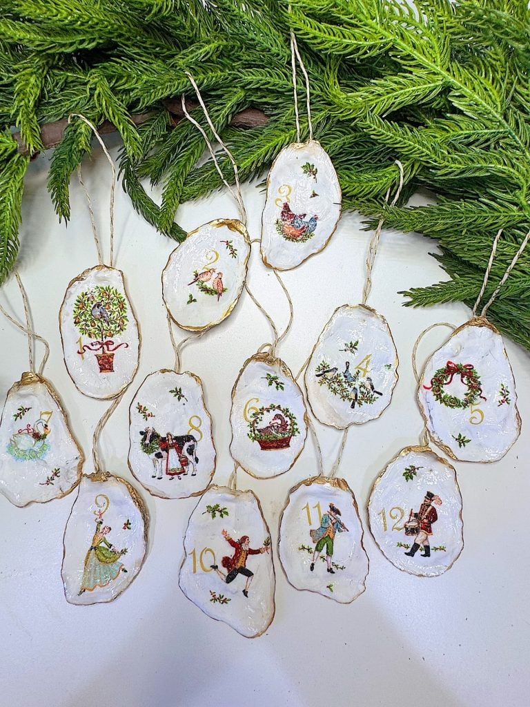 Twelve hand-painted Christmas ornaments with different holiday designs and numbers, arranged on a white surface with green pine branches at the top.