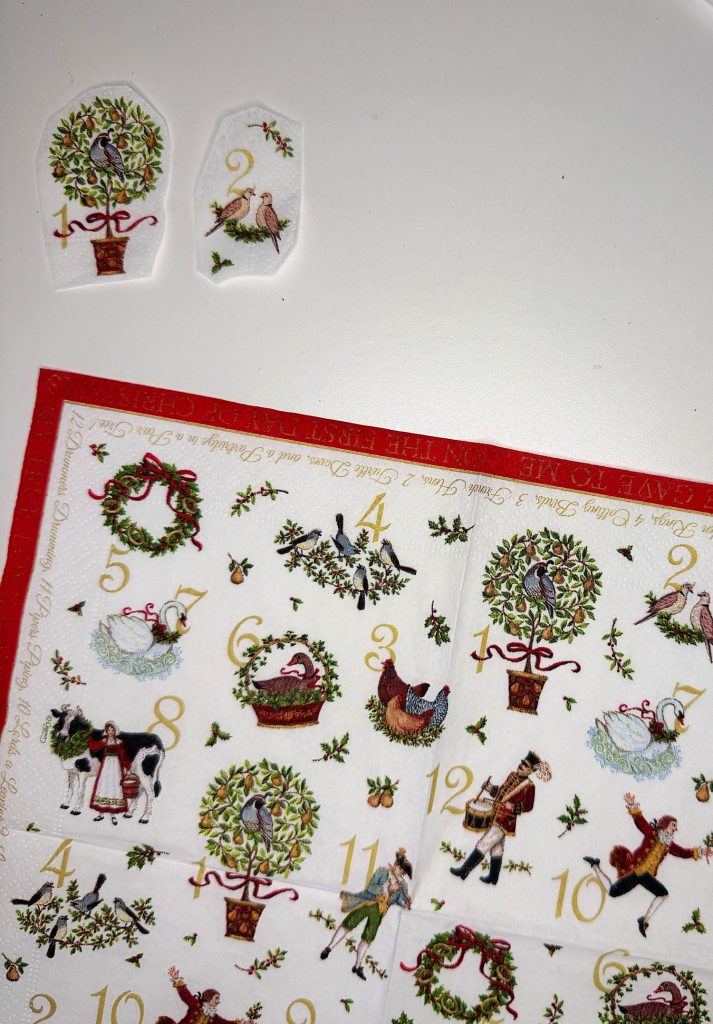 A decorative napkin with illustrations for the Twelve Days of Christmas, featuring numbers and holiday-themed images, with two sections cut out and placed above it.