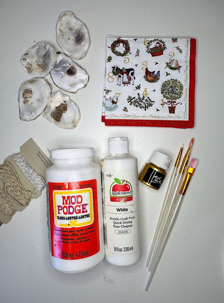 Assorted craft supplies including oyster shells, Mod Podge, white acrylic paint, gold paint, paintbrushes, twine, and a Christmas-themed napkin arranged on a white surface.