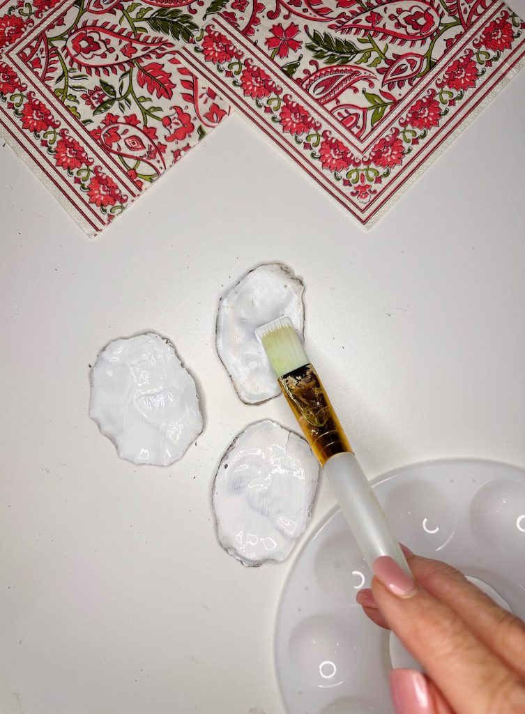 A hand holding a paintbrush applies white paint or glue to three oyster shells on a white surface, with patterned napkins in the corner.