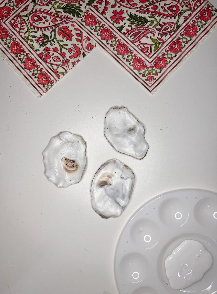 Three empty oyster shells are placed on a white surface next to a ceramic paint palette and two patterned red and green napkins.