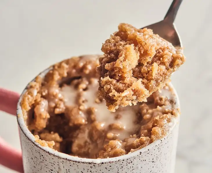 A close-up of a cinnamon-flavored mug cake with icing, with a spoon lifting a bite from the mug.