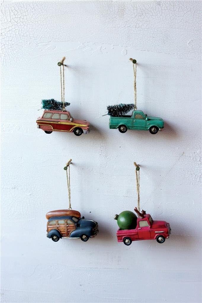 Four vintage-style car ornaments, each carrying a Christmas tree or gift, hang from strings against a white textured background.