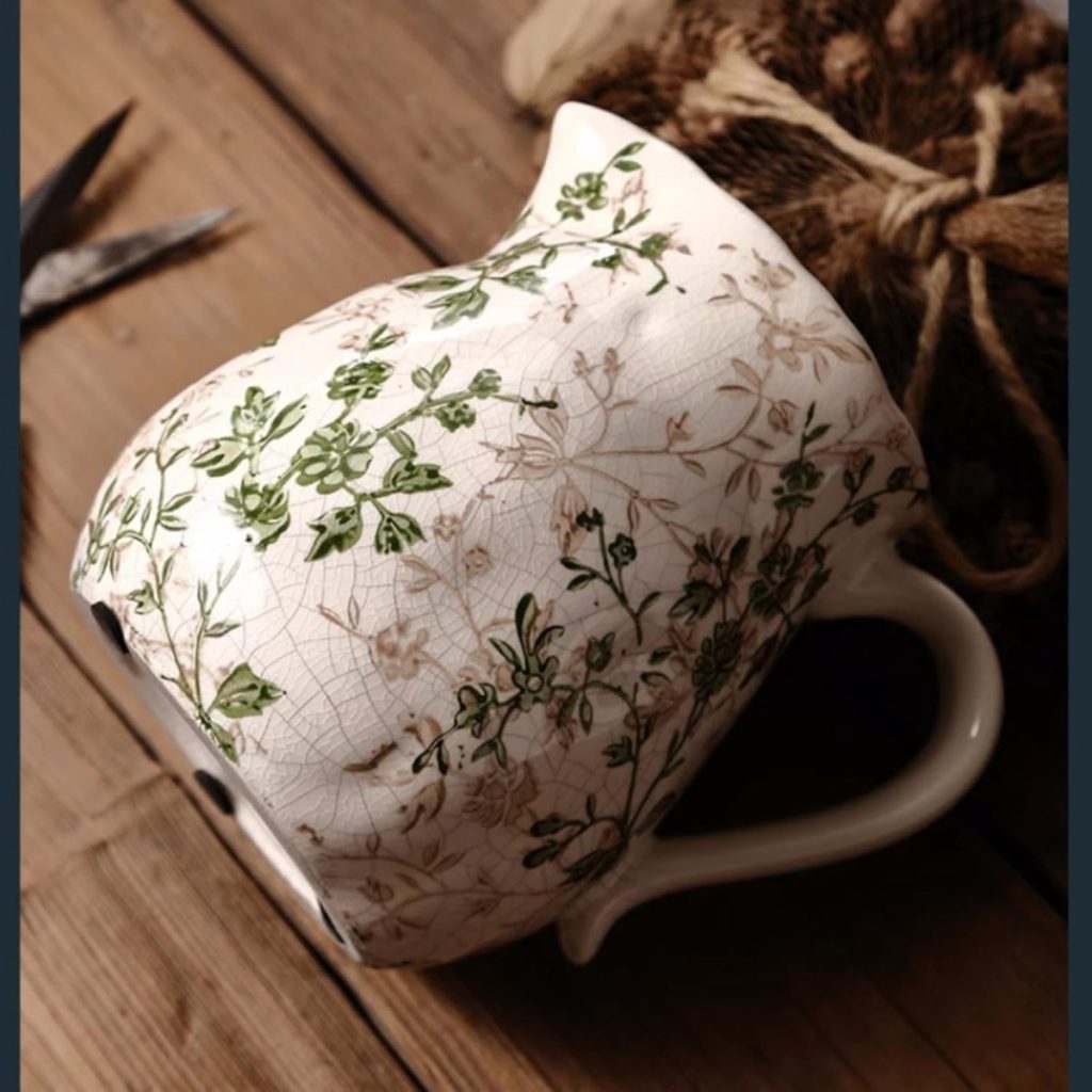 A ceramic mug with a handle, featuring a green and brown floral pattern, lies on its side on a wooden surface next to twine and scissors.
