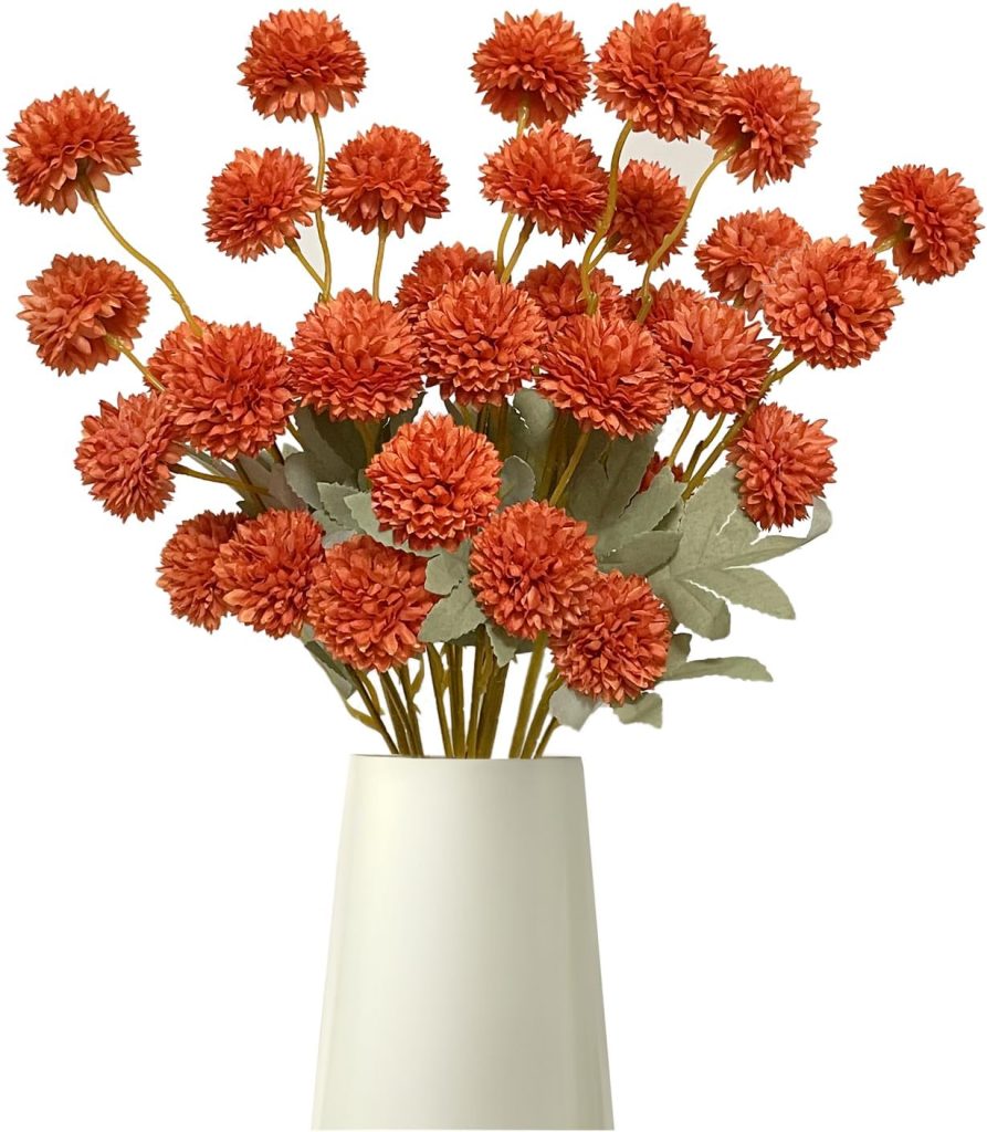 A white vase holds several stems of artificial orange pompom flowers with green leaves, arranged in a bouquet—perfect for pairing with painted pumpkins for a festive touch.
