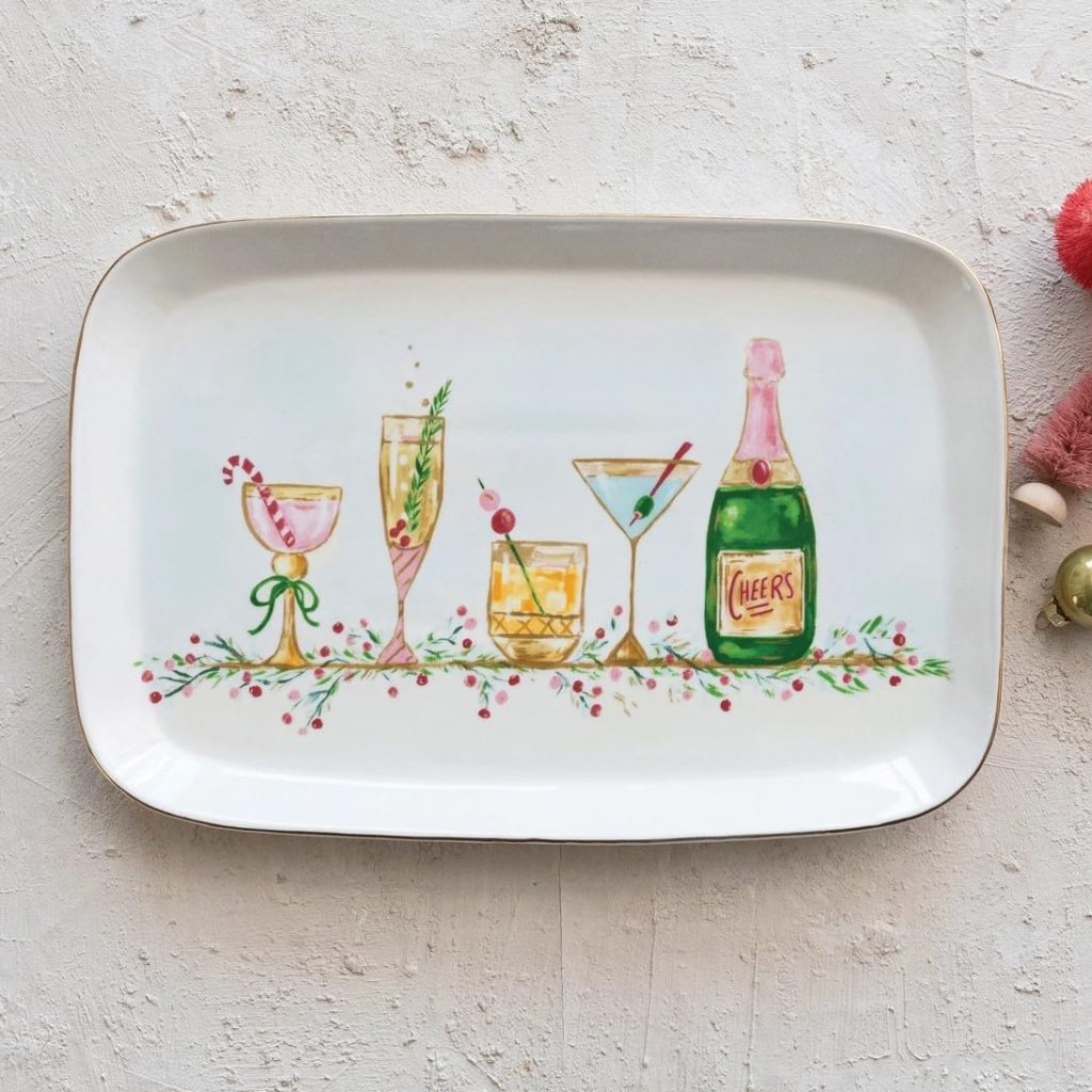 A rectangular white plate features illustrations of four festive cocktails and a bottle labeled "Cheers," decorated with holiday greenery and berries.