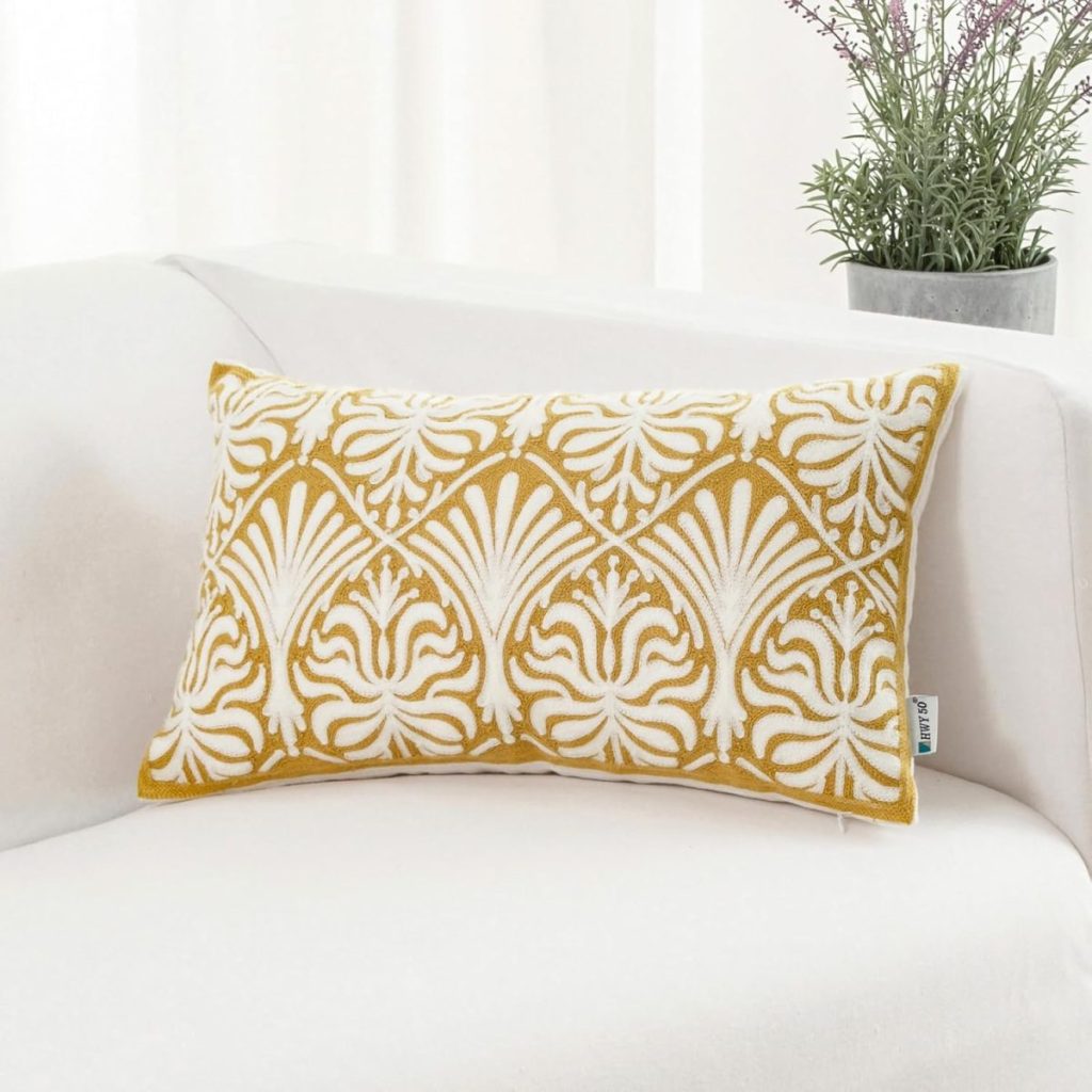 A rectangular mustard yellow and white patterned throw pillow rests on a white sofa, with a potted plant in the background.
