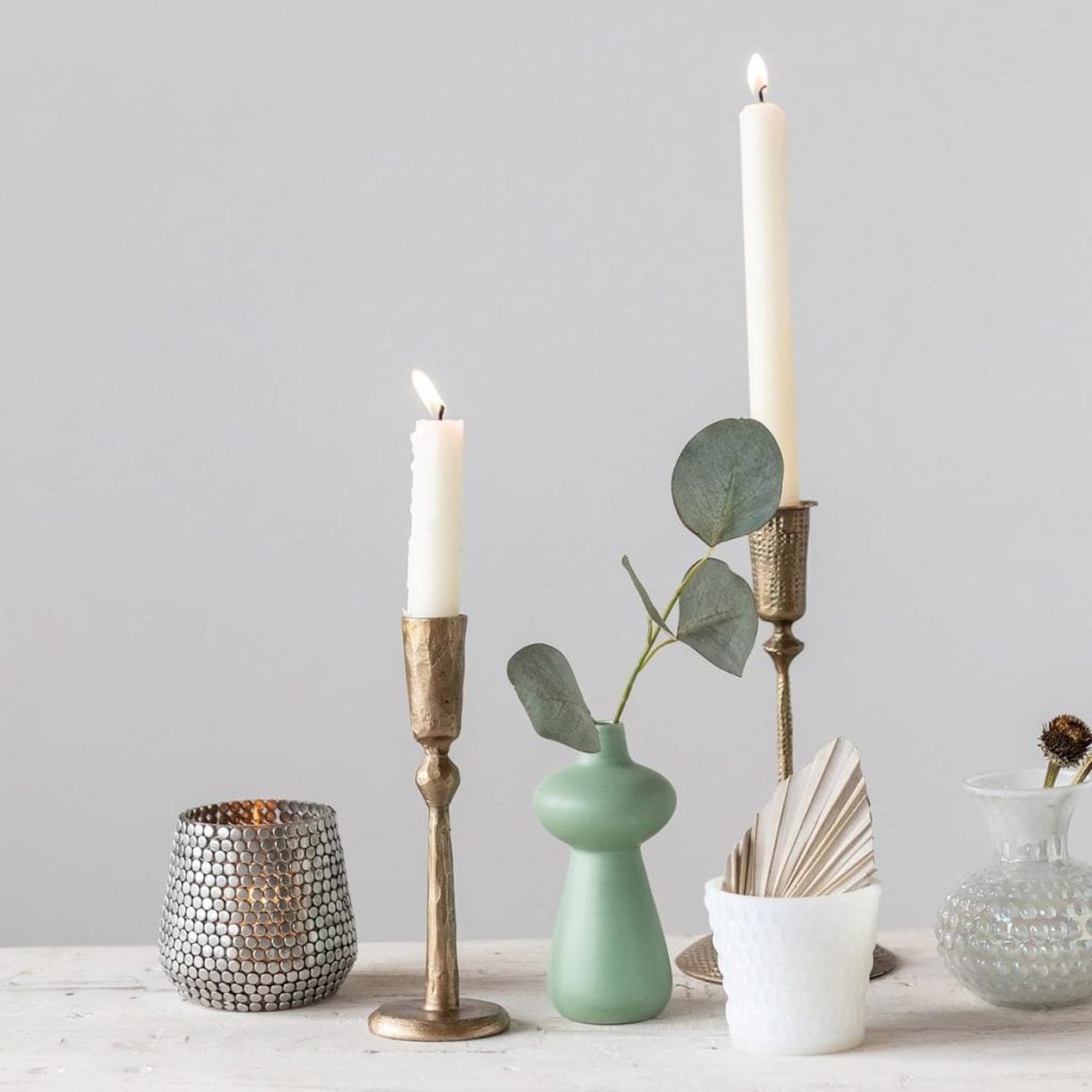 A lit white candle in a brass holder, a silver textured votive, a small green vase with eucalyptus, a white cup with folded paper, and a glass vase on a white surface.