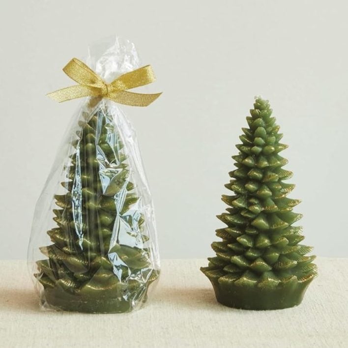 Two green, tree-shaped candles; one is wrapped in clear plastic with a gold ribbon, and the other is unwrapped, both placed on a neutral fabric surface.