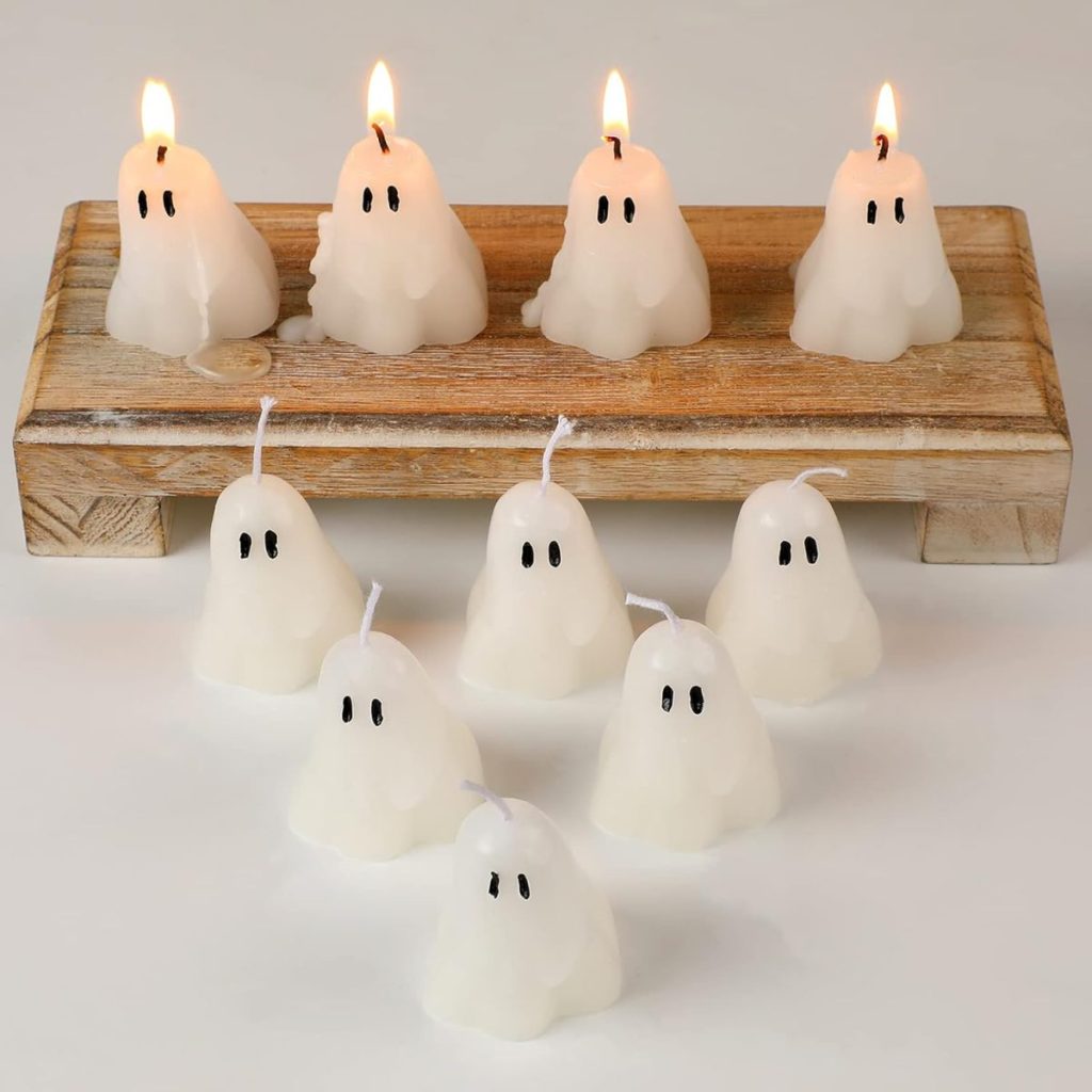 Nine ghost-shaped white candles, three of which are lit, are displayed on a wooden stand and a white surface. Each candle has two black eyes painted on it.