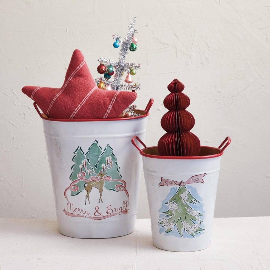Two white metal buckets with Christmas designs hold a red plaid star pillow, a small decorated tree, and a red paper ornament, against a plain light wall.