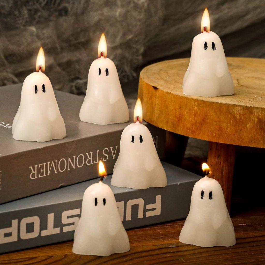Five ghost-shaped white candles with lit flames are arranged on books and a wooden surface, with a wooden stool and a gray background.