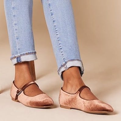 A person sits on a wooden chair, wearing light blue jeans, a white long-sleeve shirt, and pink suede Mary Jane flats—perfect style inspiration for Amazon Prime Big Deal Days shopping.