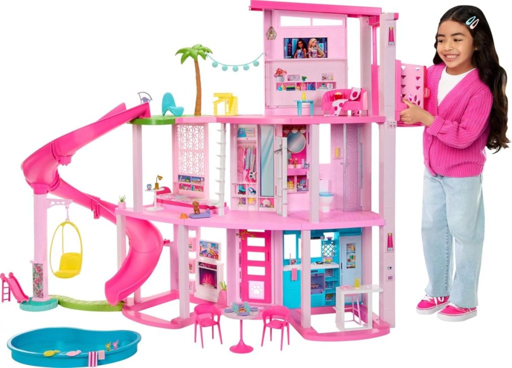 A girl stands next to a large pink dollhouse with multiple rooms, a slide, a swing, and an outdoor pool—perfect for gifting during Amazon Prime Big Deal Days.