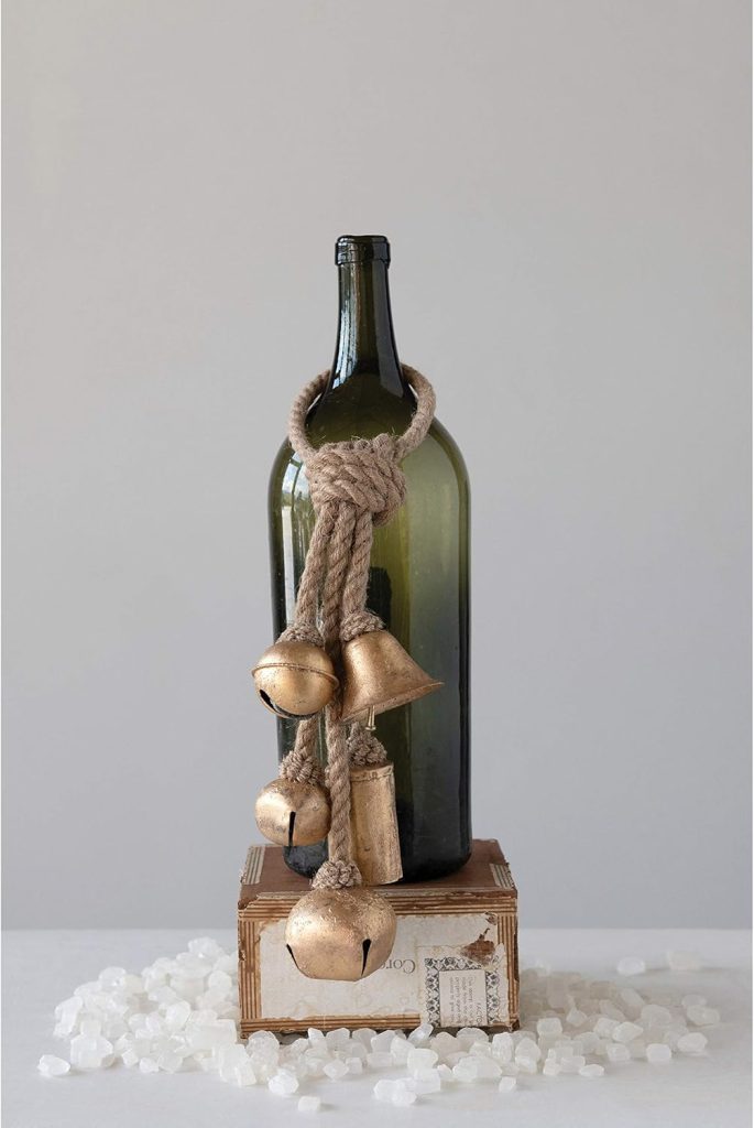 A green glass bottle with a rope and gold bells draped around its neck stands on a wooden box, surrounded by scattered white stones.