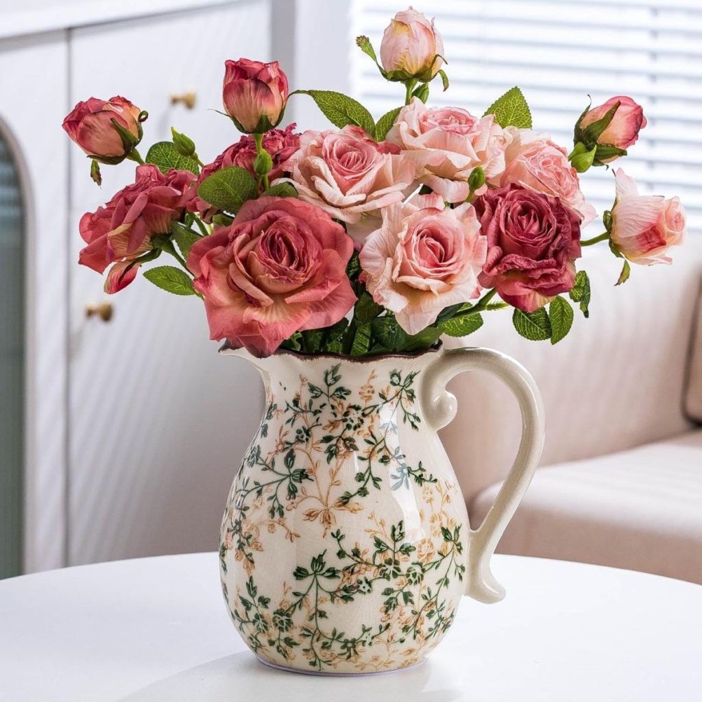 A ceramic pitcher with a floral pattern holds a bouquet of pink and blush roses on a white table in a bright room.