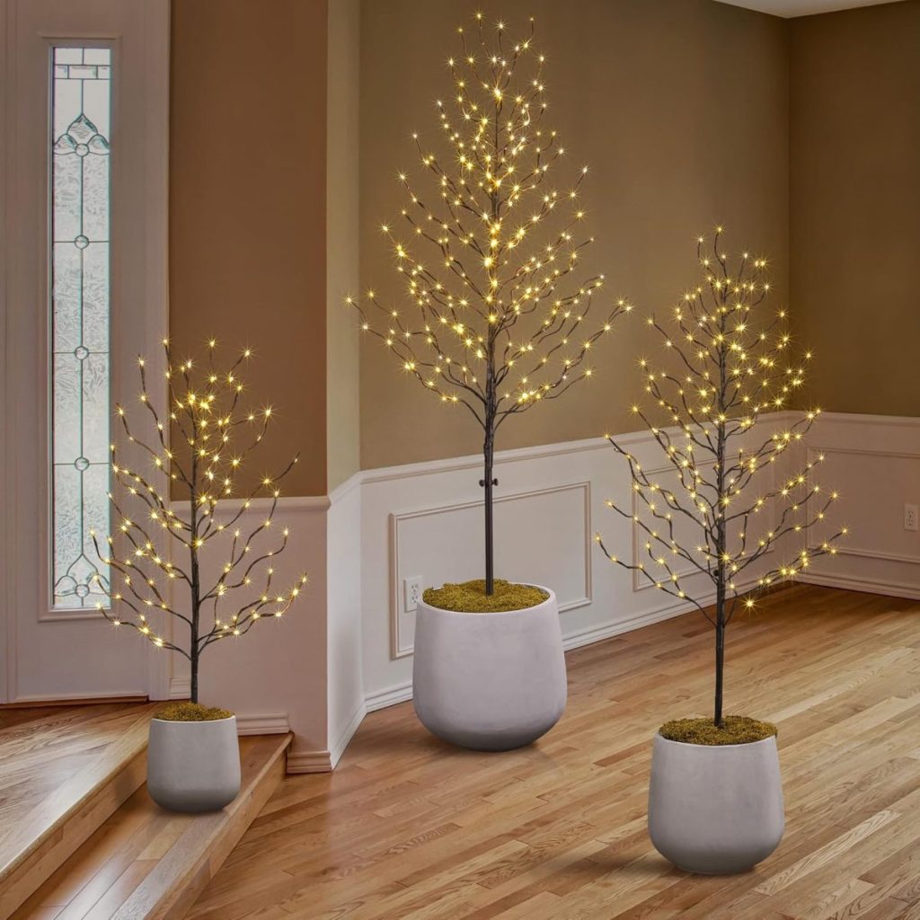 Three potted artificial trees with LED string lights add a warm glow to a corner of the room with wood flooring and beige walls—perfect decor inspiration just in time for Amazon Prime Big Deal Days.