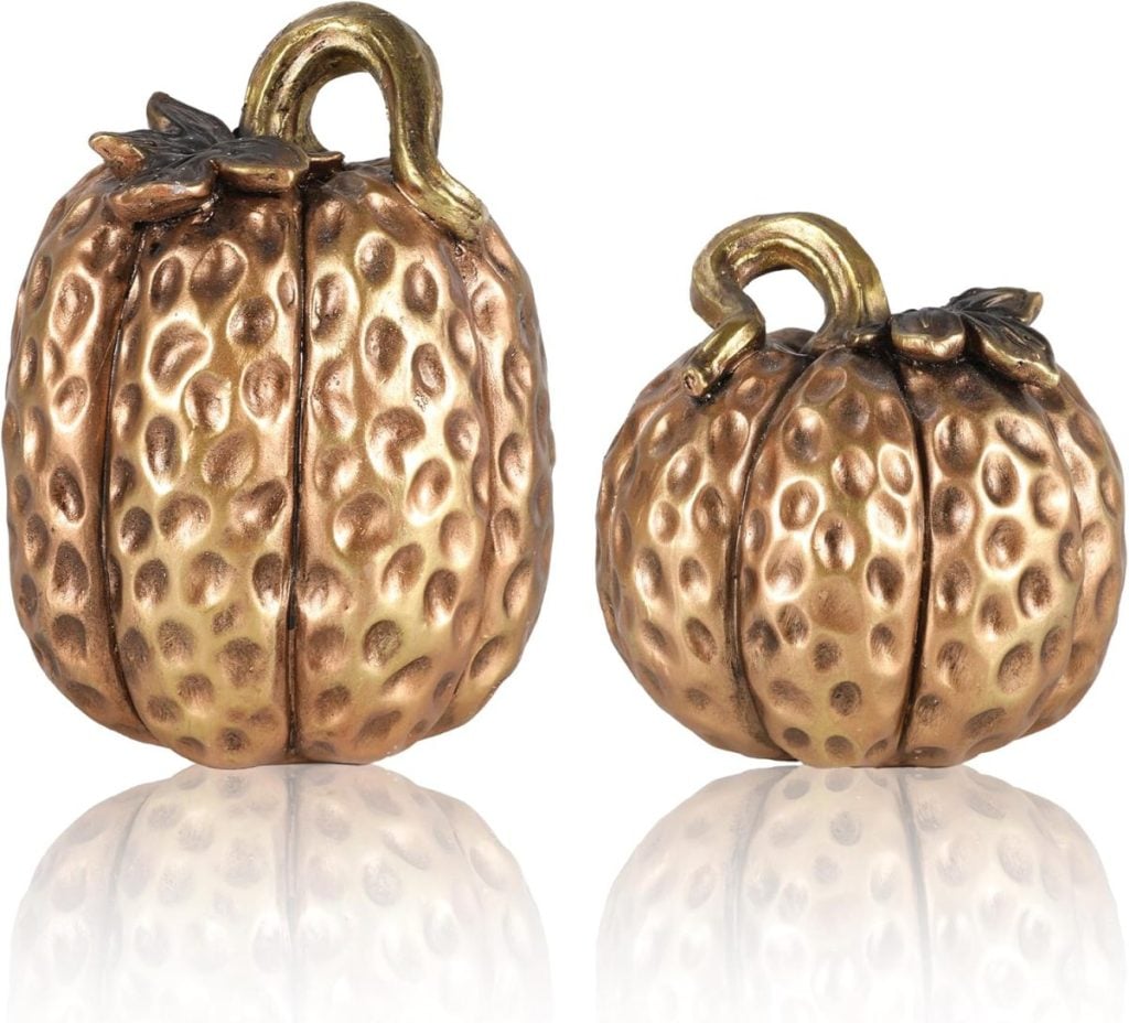 Two metallic, gold-colored decorative pumpkins with textured, bumpy surfaces and reflective bases are shown against a white background.