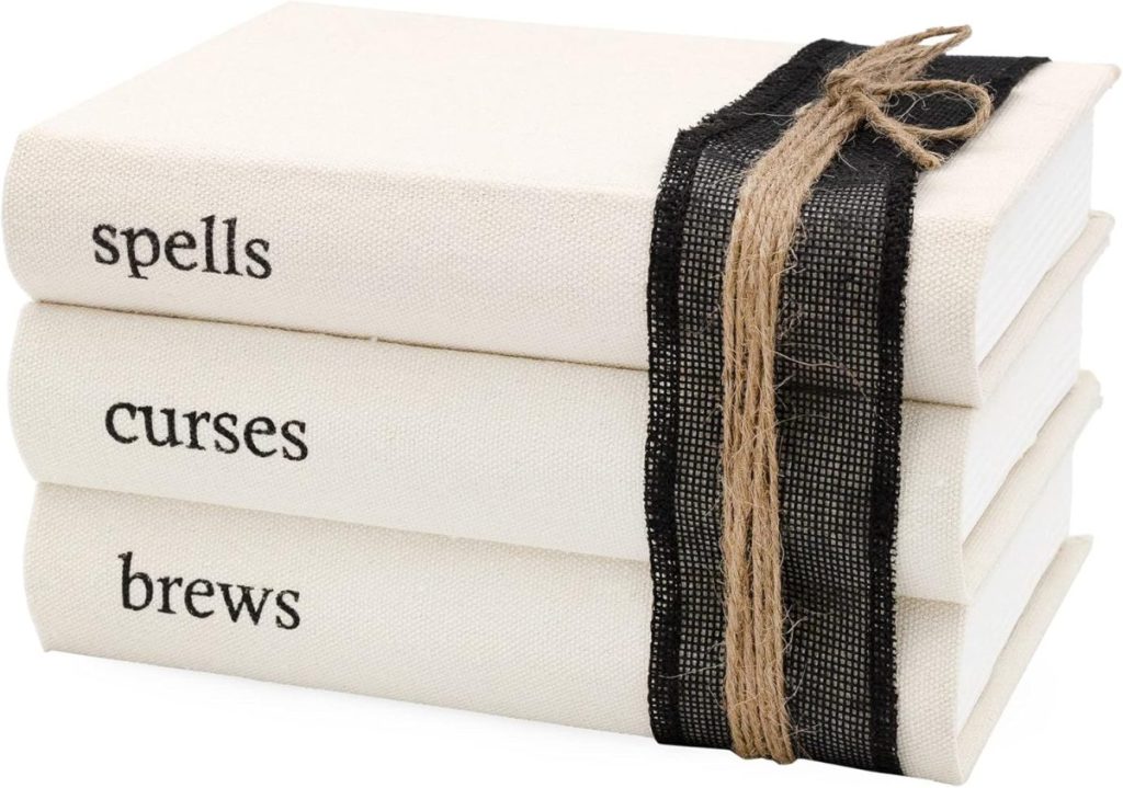 Three stacked white books labeled "spells," "curses," and "brews," tied together with black ribbon and brown twine.