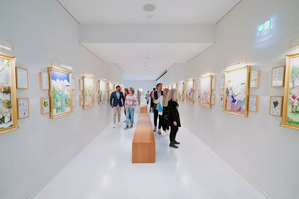 A group of people walk through a brightly lit art gallery hallway with framed paintings displayed on both walls and a long wooden bench in the center.